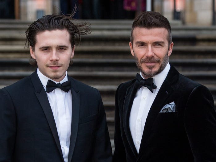 Brooklyn Beckham and David Beckham wear black suits as they're photographed on steps.